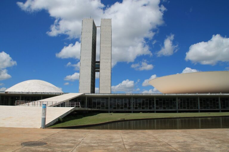 A imagem mostra o prédio do Congresso Nacional Brasileiro em Brasília, com duas torres altas, uma cúpula branca à esquerda e uma estrutura branca em forma de tigela à direita, sob um céu azul - uma visão icônica semelhante às encontradas nas interfaces de software para lotéricas.
