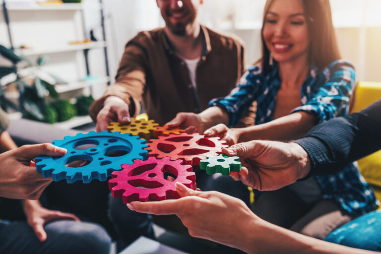 Um grupo de pessoas senta-se junto, cada uma segurando engrenagens coloridas interligadas, simbolizando o trabalho em equipe e a colaboração - muito parecido com um software para lotéricas que traz eficiência e unidade para os negócios de loteria. O foco está em suas mãos e nas engrenagens.