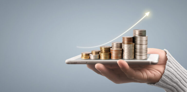 Uma mão segura um smartphone com pilhas de moedas em cima, organizadas em ordem crescente. Uma seta para cima sobre as moedas simboliza o crescimento financeiro, destacando os benefícios de um software para loterias eficiente.