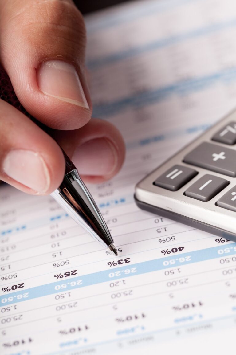 Um close-up de uma mão segurando uma caneta, apontando para dados financeiros em uma planilha impressa, com porcentagens e números visíveis. Uma calculadora é colocada ao lado do papel, ilustrando a análise com o software para lotéricas.