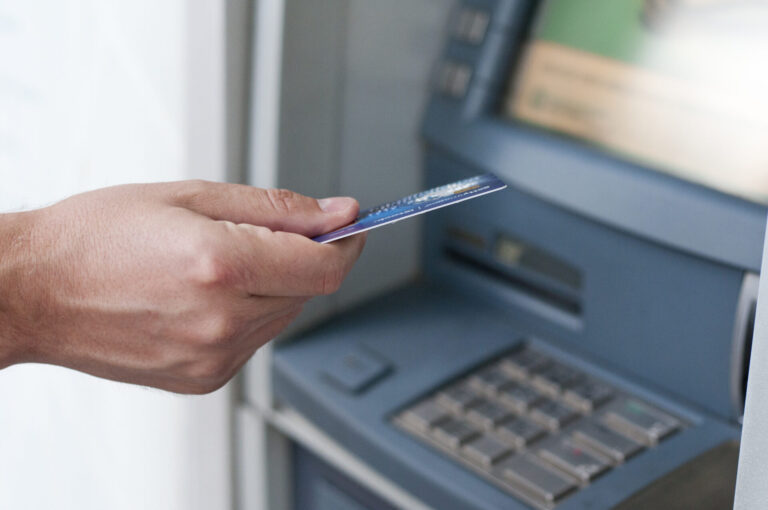 A mão de uma pessoa segura um cartão bancário em frente a um caixa eletrônico, preparando-se para inseri-lo no slot de cartão acima do teclado e da tela - ideal para quem busca um software para lotéricas moderno e eficiente.