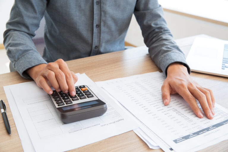 Uma pessoa de camisa cinza usa uma calculadora e analisa documentos financeiros em uma mesa, ao lado de um laptop com software para lotéricas e uma caneta.