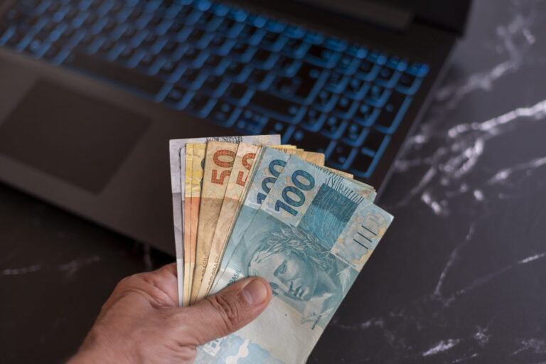 Uma mão segurando cédulas de real brasileiro de várias denominações em frente a um teclado de laptop em uma superfície de mármore preto, ilustrando transações financeiras gerenciadas com um sistema para lotéricas.