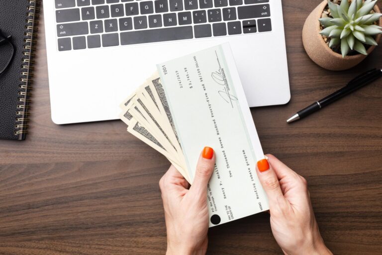 Uma pessoa com esmalte laranja segura um contracheque e notas de dólar americano sobre uma mesa de madeira com um laptop executando um software para lotéricas, ao lado de um caderno, uma caneta e um pequeno vaso de suculentas.