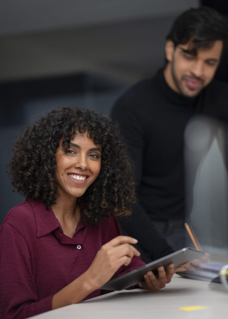 Uma mulher de cabelos cacheados, vestindo uma camisa marrom, sorri enquanto segura um tablet em uma mesa usando um sistema para lotéricas. Um homem de suéter preto está ao lado dela, olhando para baixo e escrevendo com um lápis.