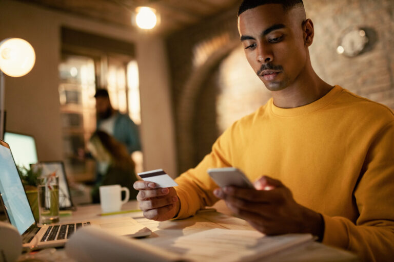 Um homem de moletom amarelo senta-se em uma mesa, concentrado em usar o software para lotéricas em seu laptop enquanto segura um cartão de crédito e um smartphone. Papéis e uma xícara de café estão sobre a mesa, com duas pessoas desfocadas ao fundo.