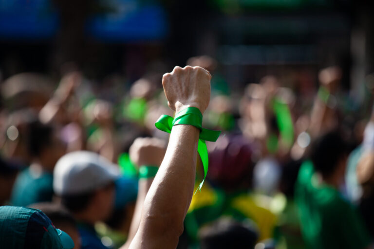 Um punho erguido envolto em uma fita verde se destaca em uma multidão de pessoas, muitas delas de verde, enquanto os lotéricos realizam novas manifestações, destacando seu papel ativo no protesto ou na manifestação.