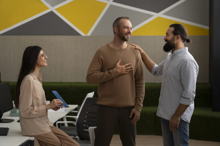 Três pessoas em um escritório, dois homens em pé e sorrindo um para o outro enquanto um dá um tapinha no ombro do outro. Uma mulher está sentada próxima com um tablet, possivelmente explorando um novo sistema para lotéricas. Decoração moderna e colorida ao fundo.