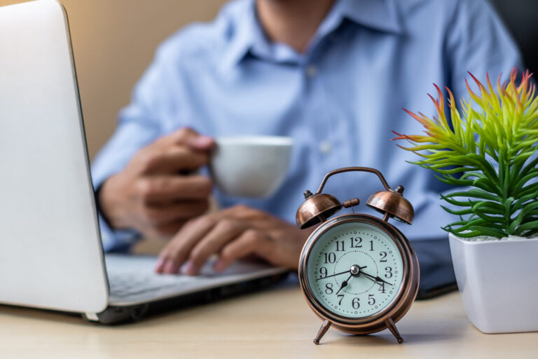 Uma pessoa de camisa azul trabalhando em um laptop e segurando uma xícara de café, com um despertador e um vaso de plantas sobre a mesa, possivelmente usando um software para lotéricas para gerenciar tarefas com eficiência.
