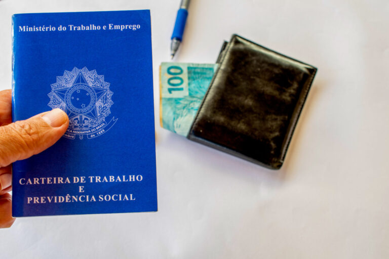 Uma mão segura uma carteira de trabalho e previdência social brasileira ao lado de uma carteira preta com uma nota de 100 reais parcialmente visível, simbolizando que os trabalhadores devem receber R 7 bilhões, e uma caneta repousa sobre a superfície branca.