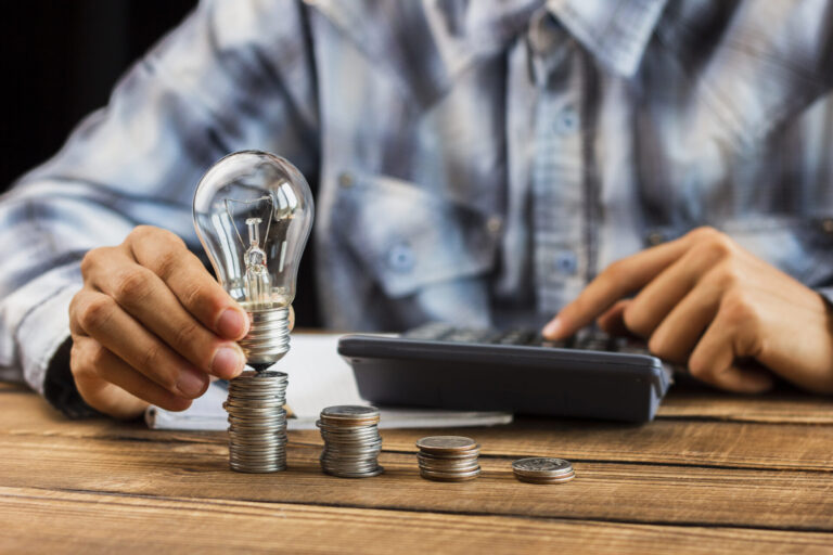 Uma pessoa de camisa xadrez segura uma lâmpada em cima de moedas empilhadas enquanto usa uma calculadora, simbolizando ideias ou inovações relacionadas a finanças - como a mansueto comenta a reformulação do marco legal sobre crescimento econômico e regulamentação.