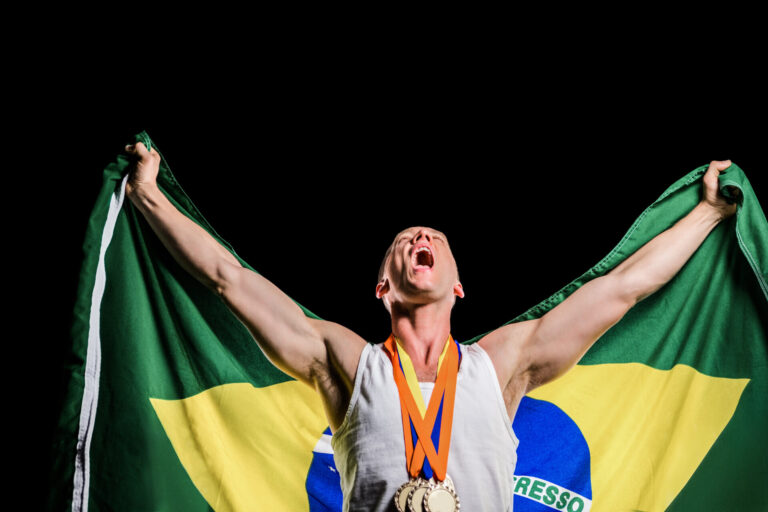 Um atleta triunfante usando medalhas levanta uma bandeira brasileira atrás de si, comemorando a vitória em um fundo preto - mostrando como as loterias caixa são uma importante fonte de apoio ao esporte nacional.