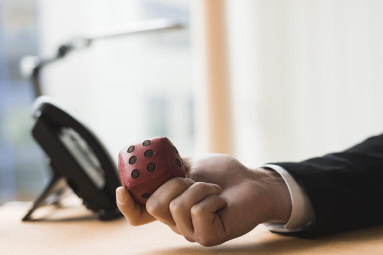 Uma pessoa de terno segura um grande dado vermelho na mão enquanto está sentada em uma escrivaninha com um telefone preto, enquanto um documento de informe febralot pl 7306/2017 está próximo contra o fundo desfocado.