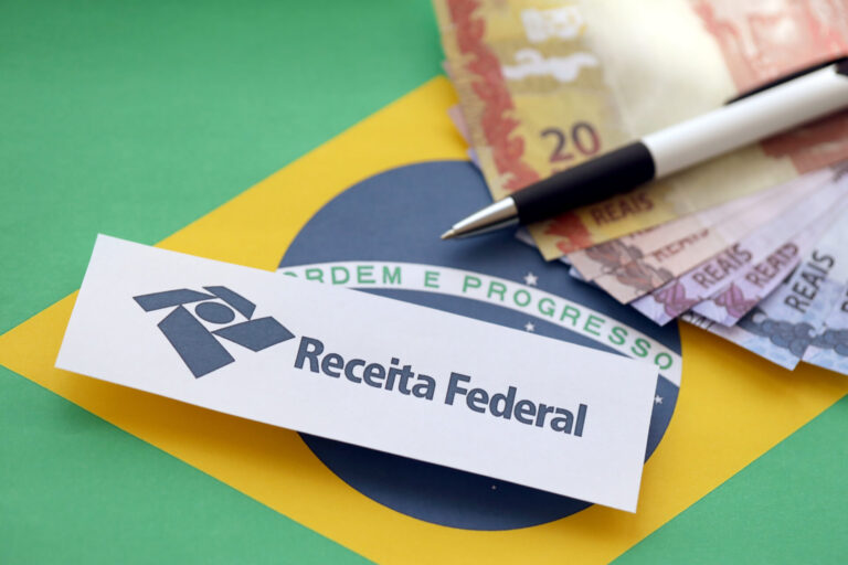 Um papel com o logotipo e o texto da Receita Federal sobre o fundo de uma bandeira brasileira, ao lado de cédulas de real e uma caneta, ilustrando como o governo aumenta o imposto no Brasil.