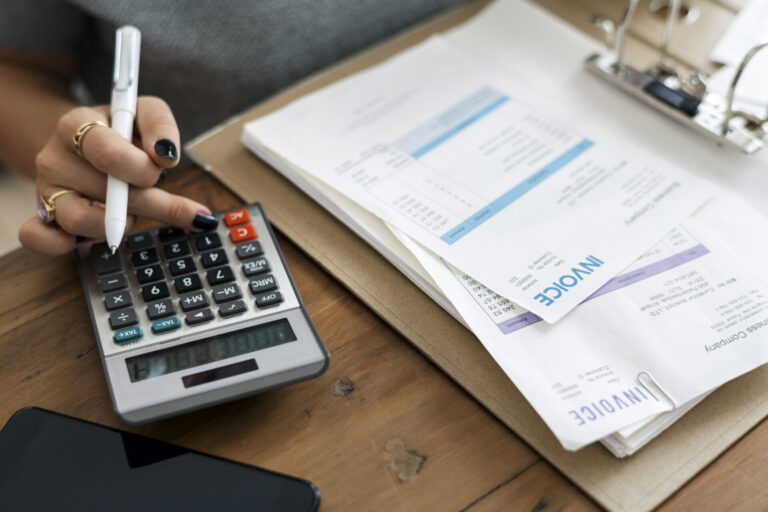 Uma pessoa usa uma calculadora enquanto segura uma caneta, com várias faturas e documentos financeiros em uma prancheta próxima em uma mesa de madeira - possivelmente analisando dados de um sistema para lotéricas. Um smartphone está parcialmente visível em primeiro plano.
