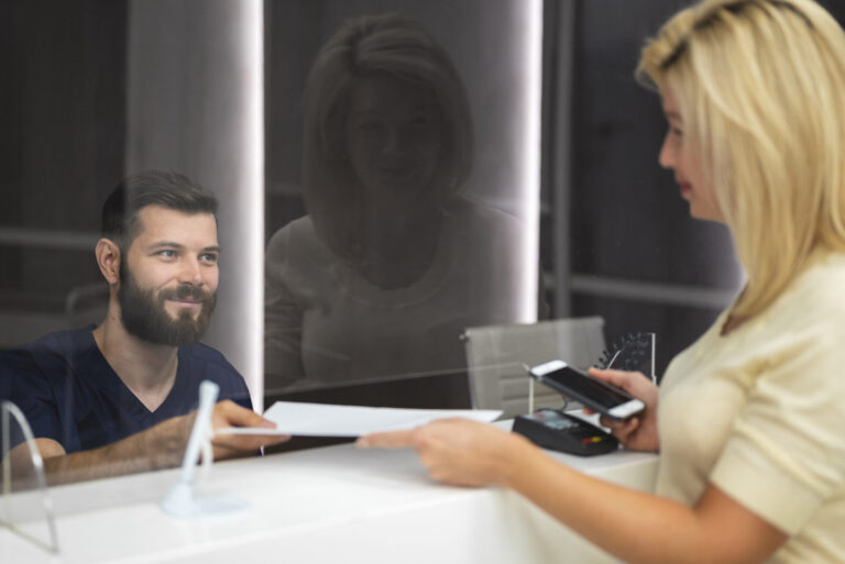Uma mulher entrega um documento a um homem barbudo atrás de um balcão com divisória de vidro, seguindo o manual operacional das atividades do correspondente caixa. Ela segura um telefone e um cartão, enquanto o homem sorri para ela do outro lado.