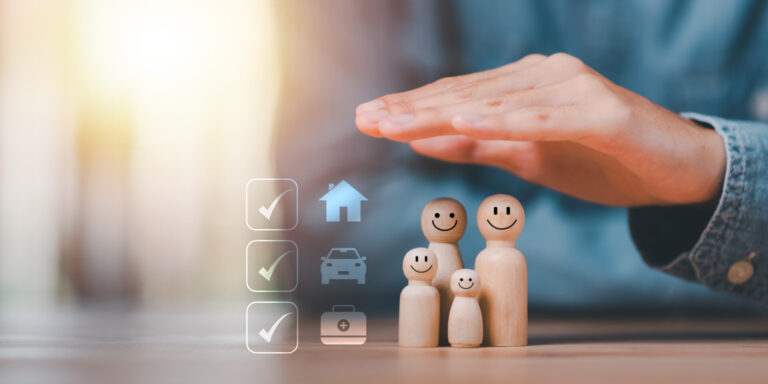 Uma mão cobre quatro figuras de madeira com rostos sorridentes, representando uma família. Ao lado deles, os ícones de uma casa, um carro e uma pasta com marcas de verificação simbolizam a proteção e o seguro de vida amparo para seus entes queridos.