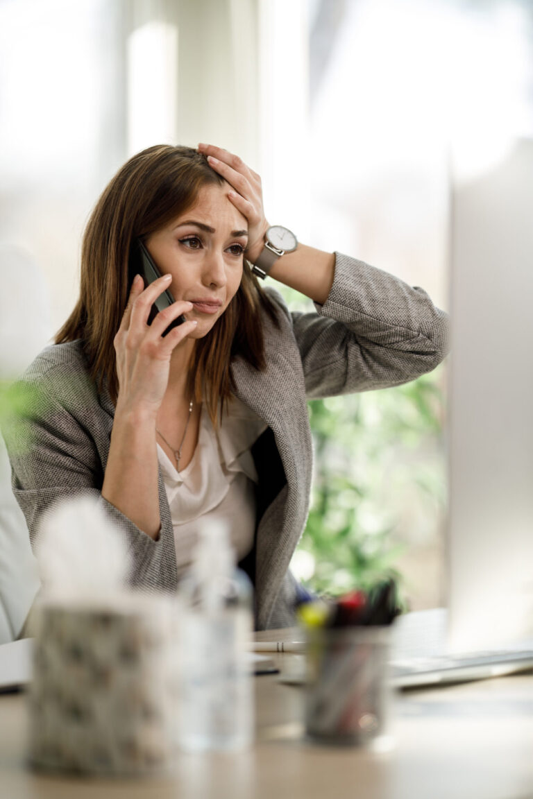 Uma mulher em traje de negócios senta-se em uma mesa, parecendo estressada enquanto fala ao telefone, possivelmente enfrentando problemas com seu software para lotéricas, com material de escritório e um computador visíveis em primeiro plano.