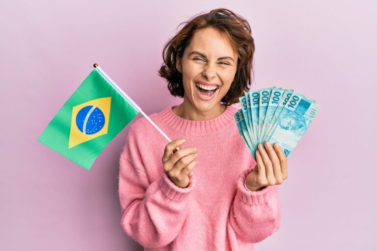 Uma pessoa sorridente com um suéter rosa segura uma bandeira do Brasil e várias notas de 100 reais, em um fundo rosa claro, comemorando com a esperança de que 2018 seja o ano da vitória.