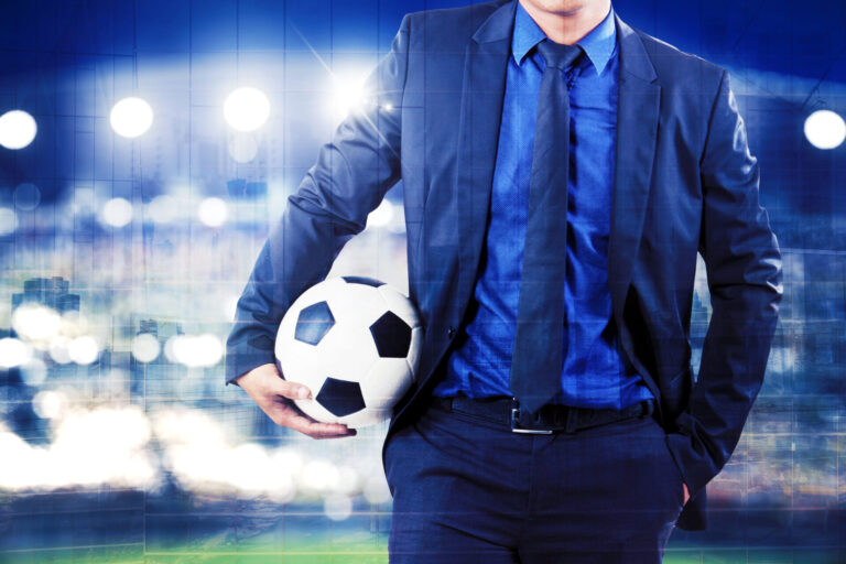 Um homem de terno azul e gravata está em um estádio à noite, segurando uma bola de futebol debaixo do braço. As luzes brilhantes do estádio brilham ao fundo, criando uma atmosfera vibrante que lembra os debates sobre a regulamentação dos jogos de azar.