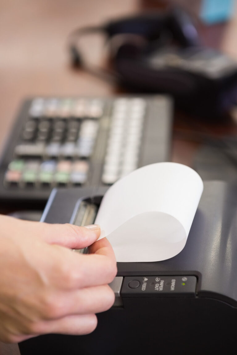 A mão de uma pessoa puxa um recibo de uma impressora térmica, com um teclado de caixa registradora embaçado ao fundo - possivelmente lidando com bobinas com miolo plástico dando problemas durante a transação.