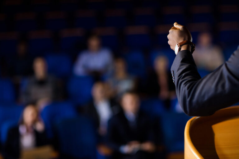 Uma pessoa de terno gesticula enquanto fala em um pódio em frente a um público desfocado sentado em cadeiras azuis, sugerindo uma apresentação formal ou palestra sobre a regulamentação dos jogos de fortuna no Brasil.