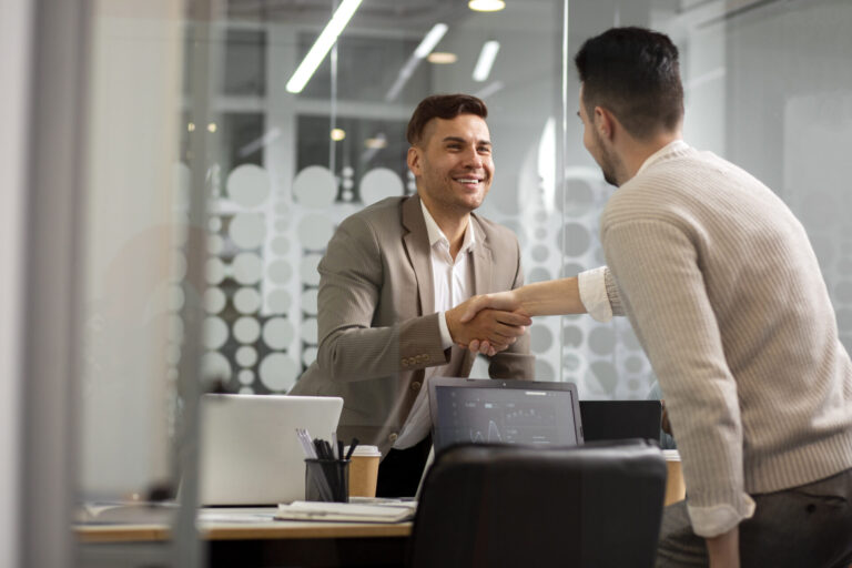 Dois homens em um escritório apertam as mãos sobre uma mesa com laptops e papéis, ambos sorrindo enquanto finalizam um acordo comercial para um software para lotéricas.