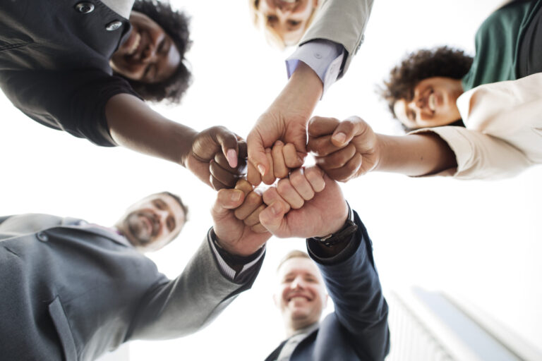 Cinco pessoas diversas em trajes de negócios estão em um círculo, sorrindo e juntando os punhos em unidade - simbolizando como os sindicatos promovem transformações no mundo empresarial, visto de baixo contra um céu claro.
