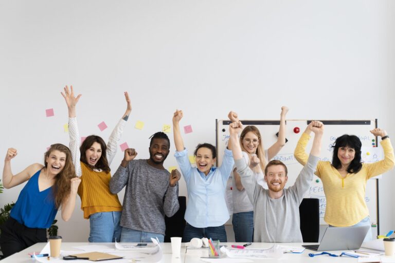 Um grupo de sete pessoas diversas e alegres está atrás de uma mesa em um escritório, sorrindo e levantando os braços em comemoração - como vencedores após ouvirem que o Dia de Sorte saiu para bolão. Papéis, notas adesivas e um quadro branco preenchem o fundo.