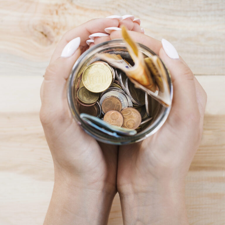 Uma pessoa com unhas bem cuidadas segura um pote de vidro cheio de moedas variadas e uma cédula dobrada - talvez ponderando se é melhor como investimento ou apenas loterias - vista de cima em uma superfície de madeira clara.