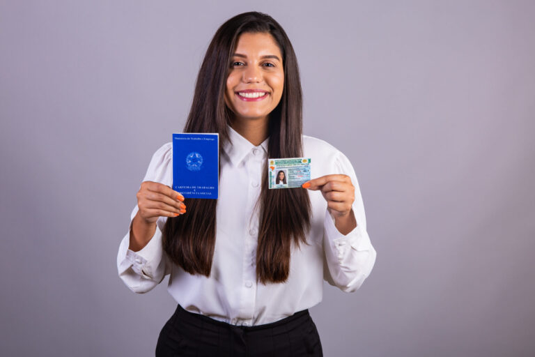 Uma mulher sorridente com longos cabelos escuros, vestindo uma blusa branca, segura um passaporte azul em uma das mãos e uma carteira de identidade na outra, ilustrando que a emissão de carteira de trabalho será cobrada contra um fundo cinza claro.