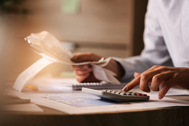 Uma pessoa revisa recibos com uma mão e usa uma calculadora com a outra, sentada em uma mesa com papéis e documentos financeiros, garantindo a conformidade com as normas legais de contabilidade durante o trabalho de orçamento ou contabilidade.