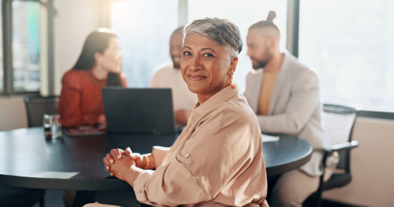 Uma mulher idosa sorridente, com cabelos grisalhos curtos, senta-se em uma mesa redonda em um escritório bem iluminado, supervisionando atividades operacionais gerenciais enquanto três colegas ao fundo conversam.