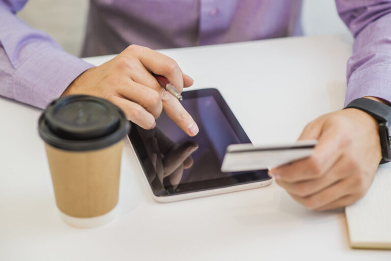 Uma pessoa usa um tablet com uma mão enquanto segura um cartão de crédito com a outra, possivelmente navegando em sites como projetos coíbem o uso de jogos de azar. Uma xícara de café e um notebook estão sobre a mesa branca; a pessoa veste uma camisa roxa e usa relógio.