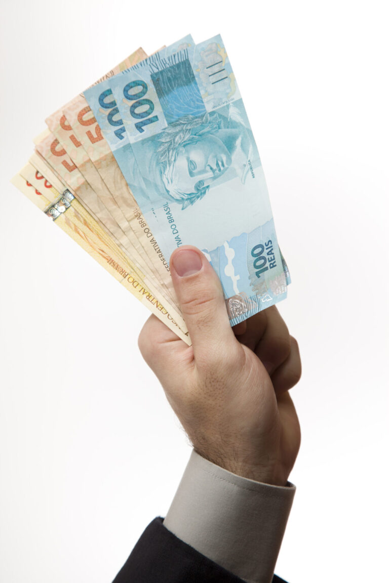 Uma mão segurando várias cédulas de real de diferentes denominações, incluindo notas de 100 e 50 reais bem visíveis, frequentemente vistas no contexto de uma distribuição dos prêmios de loterias, contra um fundo branco.