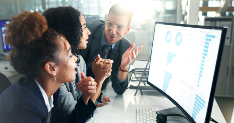 Três colegas em trajes de trabalho discutem dados em um monitor de computador que mostra tabelas e gráficos, destacando aspectos do treinamento de pessoal: do planejamento à implementação, em um ambiente de escritório moderno e bem iluminado.