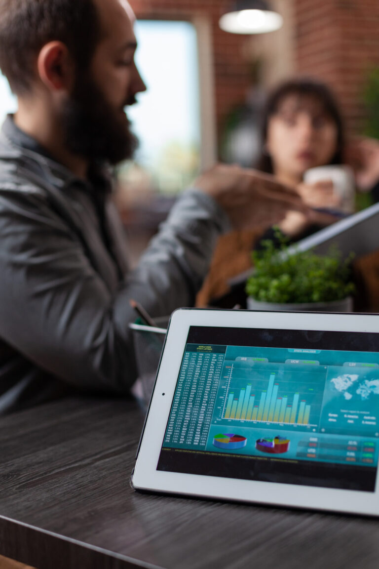 Um tablet em uma mesa exibe tabelas e gráficos financeiros coloridos, sugerindo por que fazer a transição de planilhas, enquanto duas pessoas discutem em um ambiente de escritório moderno. Uma pequena planta fica ao lado do tablet.