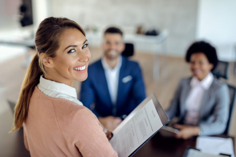 Uma mulher sorridente segurando um currículo olha para a câmera, enquanto duas pessoas vestidas profissionalmente sentam-se em frente a ela em uma mesa, sugerindo uma entrevista de emprego focada em suas habilidades para prestar um bom atendimento ao cliente em um ambiente de escritório.