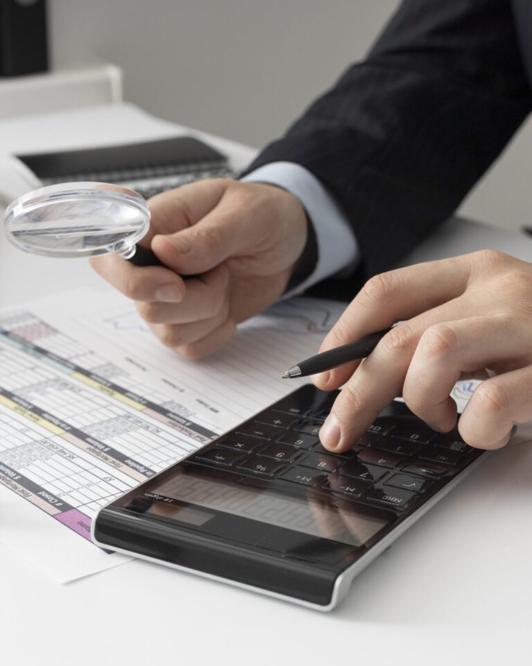 Uma pessoa de terno usa uma calculadora e segura uma caneta e uma lupa, analisando documentos financeiros com tabelas impressas - demonstrando como colocar a gestão financeira no dia a dia de forma eficaz.
