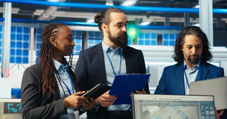 Três engenheiros em trajes de negócios discutem o trabalho em um escritório moderno. Um segura uma prancheta, outro um tablet e o terceiro trabalha em um laptop, analisando como a Caixa define novos VPs e funde loterias enquanto diagramas técnicos aparecem nas telas próximas.