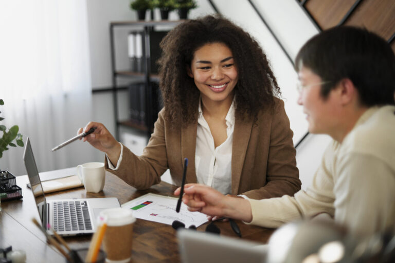 Duas pessoas sentadas em uma mesa com laptops e documentos, sorrindo e conversando sobre trabalho. Compartilhando dicas para deixar sua equipe mais produtiva, um gesticula com uma caneta enquanto o outro ouve em um escritório moderno e iluminado com plantas e prateleiras.