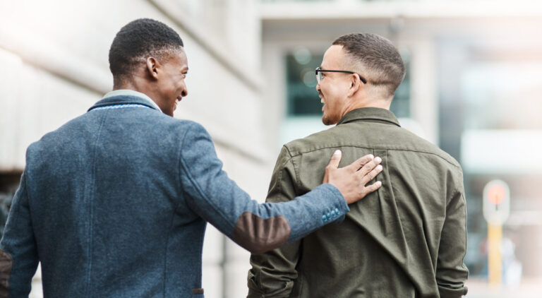 Dois homens caminhando ao ar livre em um ambiente urbano, sorrindo um para o outro enquanto discutem como abordar clientes da lotérica. Um homem tem a mão nas costas do outro em um gesto amigável, sugerindo uma conversa agradável e de apoio.