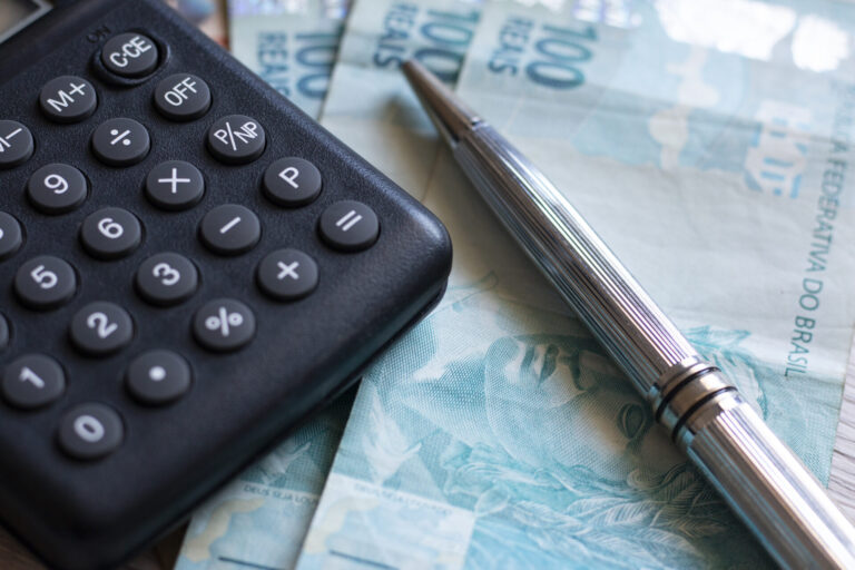 Um close-up de uma calculadora preta, uma caneta prateada e várias notas de 100 reais em uma superfície - perfeito para ilustrar o balanced scorecard o que é e como é aplicado nas finanças.