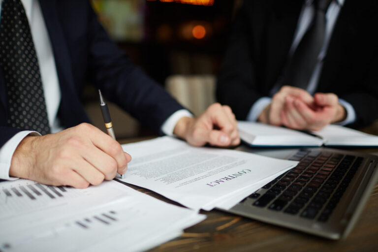 Duas pessoas em ternos de negócios sentam-se em uma mesa, analisando documentos rotulados como "CONTRATO", enquanto um laptop e papéis com gráficos sugerem que a comissão discute a regulamentação dos jogos on-line. Uma pessoa segura uma caneta, pronta para escrever ou assinar.