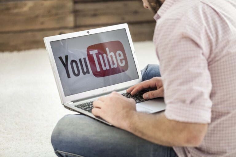 Uma pessoa vestindo uma camisa xadrez rosa está sentada no chão usando um laptop com o logotipo do YouTube exibido, navegando em vídeos lotéricos.
