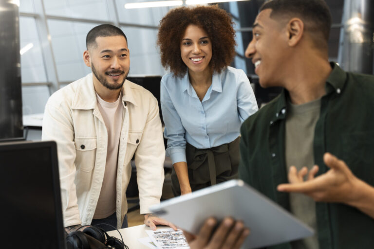 Três jovens profissionais, personificando as fases do empreendedorismo lotérico, reúnem-se em um escritório moderno. Um homem segura um tablet enquanto fala e os outros ouvem e sorriem, envolvidos em sua discussão em meio aos monitores de computador nas mesas.