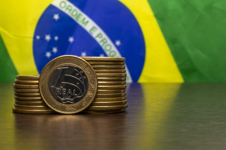 Pilhas de moedas de real são colocadas em uma superfície de madeira com uma bandeira do Brasil ao fundo, enquanto o deputado vermelho afirma que o país está enfrentando desafios econômicos.