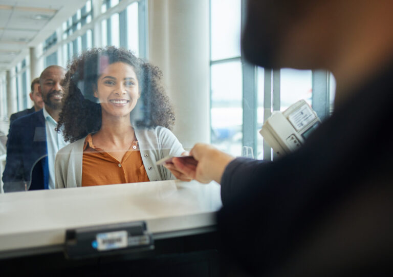 Uma mulher sorri enquanto entrega seu passaporte a um funcionário do aeroporto em um balcão de check-in, com um homem atrás dela na fila, enquanto as notícias sobre o presidente da Caixa confirmando o reajuste das apostas passam em uma TV próxima.
