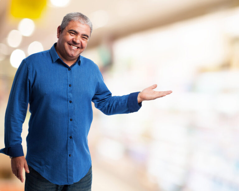 Um homem sorridente de camisa azul está em um ambiente interno, estendendo o braço direito como se estivesse apresentando ou convidando a atenção para algo ao lado - talvez destacando seu papel lotérico fundamental. O fundo está suavemente desfocado.