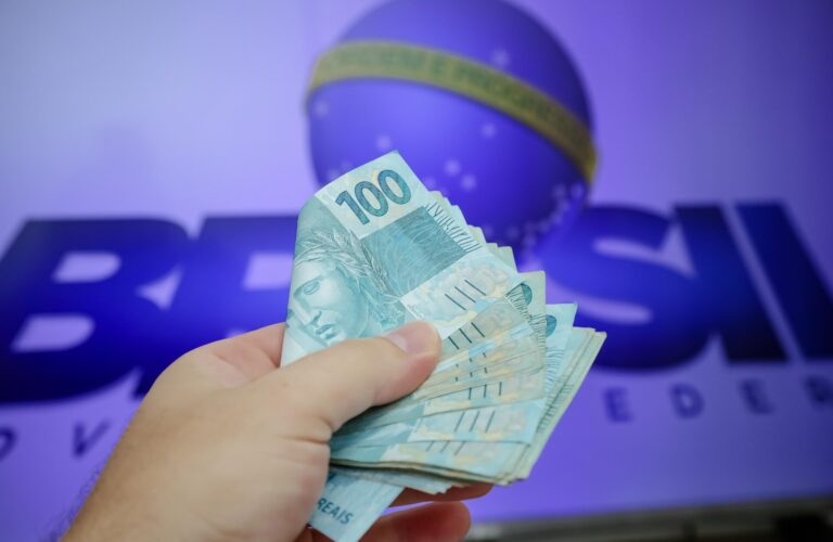 Uma mão segura várias cédulas de 100 reais em frente a um fundo desfocado com o logotipo da Caixa Econômica Federal e um globo com uma faixa da bandeira brasileira, ilustrando como a abertura do mercado justificou reajuste para o governo.
